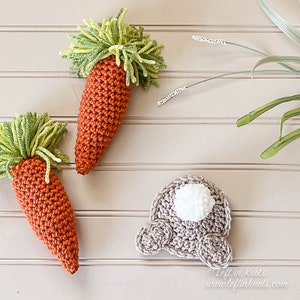 Puede incluir: Dos zanahorias de crochet con tapas verdes y un conejito de crochet con una cola de pomp&oacute;n blanco. Las zanahorias y el conejito est&aacute;n sobre una superficie de madera clara.