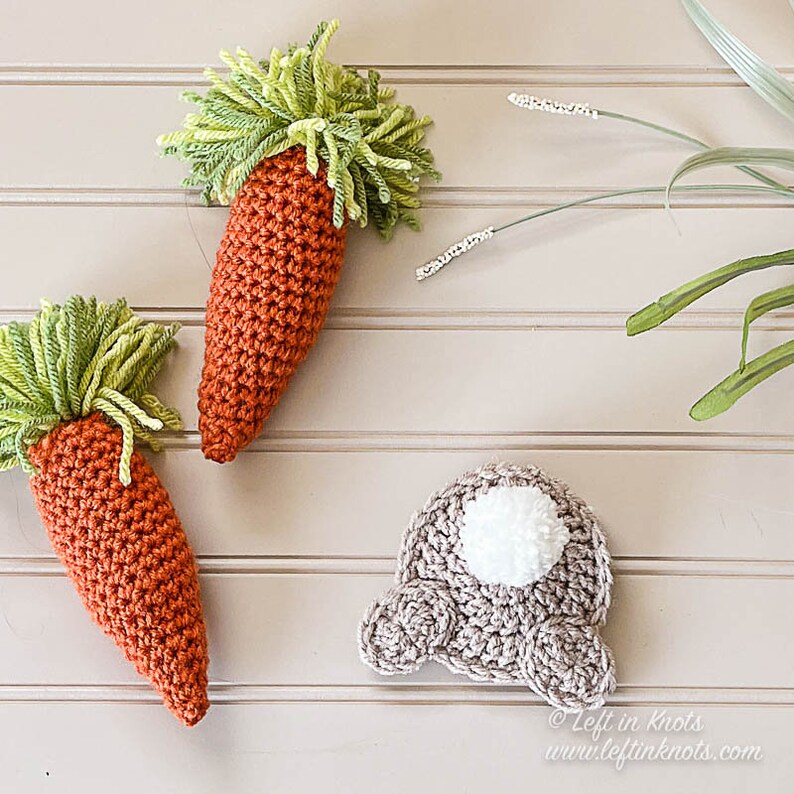 Puede incluir: Dos zanahorias de crochet con tapas verdes y un conejito de crochet con una cola de pomp&oacute;n blanco. Las zanahorias y el conejito est&aacute;n sobre una superficie de madera clara.