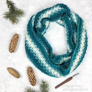 May include: A teal and white crocheted infinity scarf with a textured stitch pattern. The scarf is lying on a white surface with pine branches and pine cones.