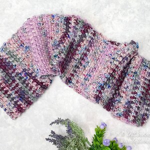 May include: A long, crocheted scarf in shades of pink, purple, and gray. The scarf is folded in half and laid flat on a white surface.
