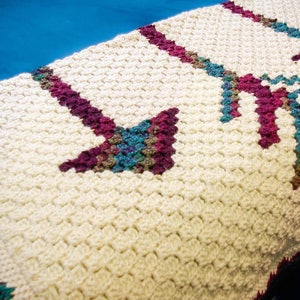 May include: A white crocheted blanket with a repeating pattern of arrows in shades of purple, teal, and brown.