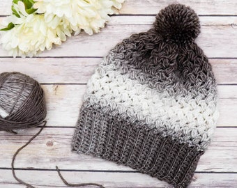 Puede incluir: Un gorro de punto con un pompón marrón. El gorro está hecho con un hilo degradado en tonos marrón, blanco y gris. El gorro está sobre una superficie de madera.
