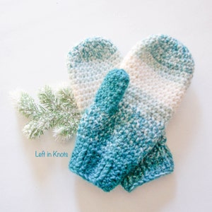 May include: A pair of teal and white crocheted mittens with a gradient pattern. The mittens are laying on a white surface with a sprig of greenery.