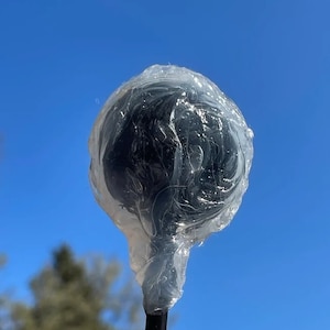 以下が含まれることがあります： 鮮やかな青い空を背景に、透明なセロファンで包まれた黒いロリポップ。ロリポップは、暗く丸いキャンディーの中心と黒い棒を持っています。セロファンはしわくちゃで、画像は屋外で撮影されています。