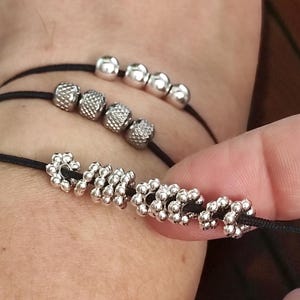 May include: Three black cord bracelets with silver beads. The bracelets feature different bead designs, including smooth spheres, textured cylinders, and floral-shaped beads. The bracelets are displayed on a wrist.