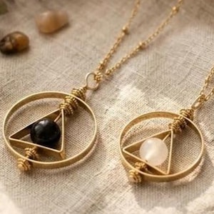 May include: Two gold-colored pendant necklaces. Each necklace features a circular frame with a triangle inside, holding a gemstone. One gemstone is black, the other is white. The necklaces are on a beige fabric.