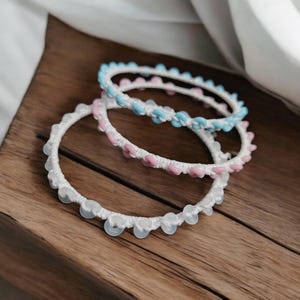 May include: Three handmade bracelets in a stack. The bracelets are white with light blue, pink, and clear accents. The bracelets are on a wooden surface with a white fabric in the background. The bracelets are a delicate and feminine accessory.