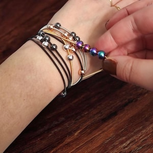 Op de afbeelding: Een pols met meerdere armbanden, waaronder een zwarte elastische armband met zilveren kralen, een gouden armband met roségouden kralen en een armband met iriserende paarse en blauwe kralen.