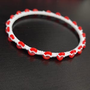 May include: A white bracelet with red beads evenly spaced around the circumference. The beads are a vibrant red color, and the bracelet appears to be handmade. The bracelet is on a dark surface.