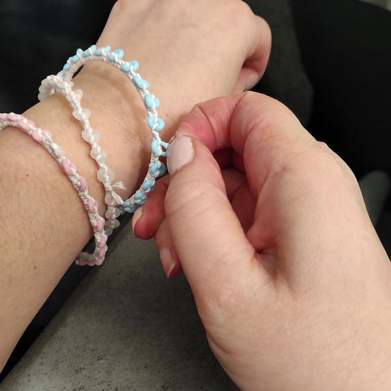 May include: Three handmade bracelets in shades of pink, white, and light blue. The bracelets are made with a woven cord and small, heart-shaped beads. One bracelet is being adjusted on a wrist.