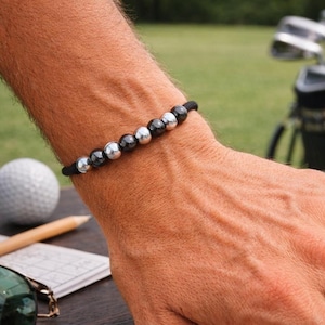 Puede incluir: Una pulsera de cuentas negras con cuentas plateadas y grises oscuras alternadas. La pulsera está ensartada en un cordón negro y se lleva en la muñeca. Una pelota de golf y un lápiz están en el fondo.