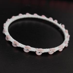 May include: A white bracelet with pink circular beads evenly spaced around the circumference. The beads are attached to a white woven band. The bracelet is set against a dark background.