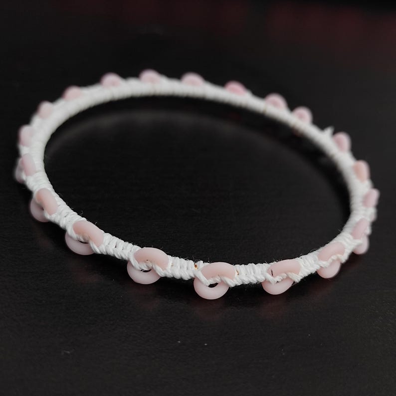 May include: A white bracelet with pink circular beads evenly spaced around the circumference. The beads are attached to a white woven band. The bracelet is set against a dark background.