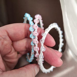 May include: Three handmade bracelets in pastel colors are held in a hand. One bracelet is light blue, one is pink, and the third is white. Each bracelet is made with small, round beads strung on white cord.