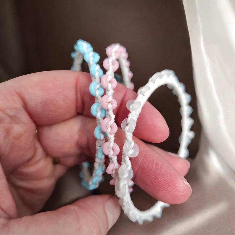 May include: Three handmade bracelets in pastel colors are held in a hand. One bracelet is light blue, one is pink, and the third is white. Each bracelet is made with small, round beads strung on white cord.