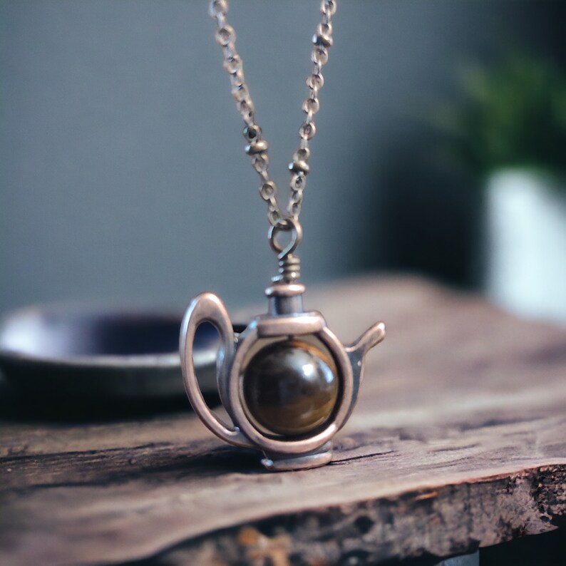 May include: A gold-toned necklace with a teapot pendant featuring a round, brown tiger's eye stone.