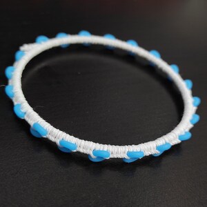 May include: A white bracelet with a woven design, adorned with evenly spaced, light blue, oval-shaped beads. The bracelet is set against a dark background, highlighting its bright colors and intricate details.
