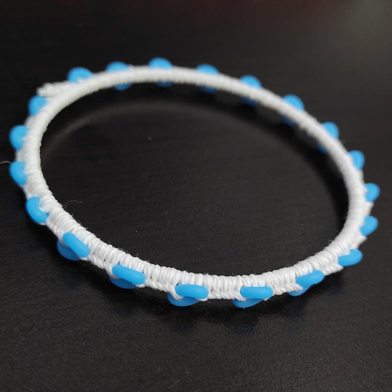 May include: A white bracelet with a woven design, adorned with evenly spaced, light blue, oval-shaped beads. The bracelet is set against a dark background, highlighting its bright colors and intricate details.