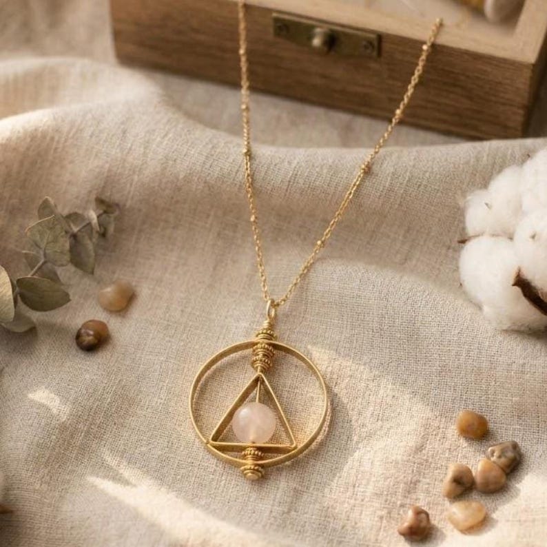 May include: A gold-colored necklace with a pendant featuring a circle and a triangle, with a pink gemstone at the center. The necklace is on a beige fabric surface, with small stones and a wooden box in the background. The necklace is a delicate piece of jewelry.
