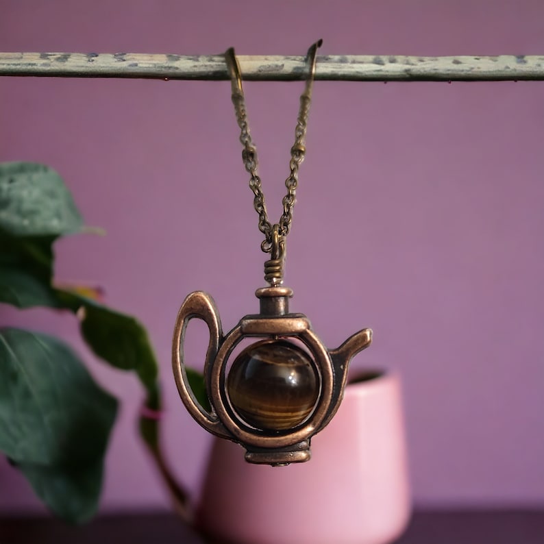 May include: A gold-toned necklace with a teapot pendant featuring a round, brown tiger's eye stone.