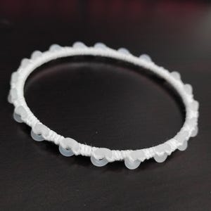 May include: White beaded bracelet. The bracelet features a white woven band adorned with small, round, translucent beads. The beads are evenly spaced around the circumference of the bracelet. The bracelet is set against a dark background.