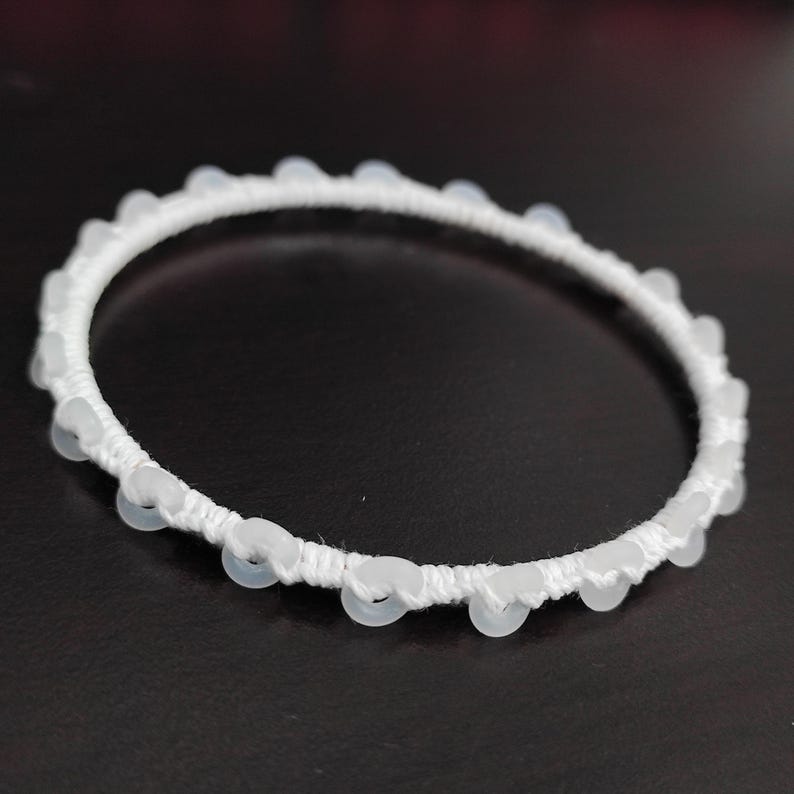 May include: White beaded bracelet. The bracelet features a white woven band adorned with small, round, translucent beads. The beads are evenly spaced around the circumference of the bracelet. The bracelet is set against a dark background.