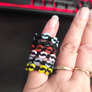 May include: A selection of colourful, linked rings in shades of blue, red, pink, and yellow, stacked on a finger. The rings are made of a glossy material and are connected by black string. A gold ring with small diamonds is also visible.