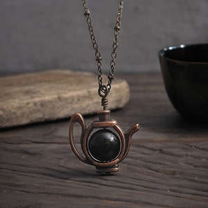 May include: A copper-toned necklace with a teapot pendant. The teapot is open and has a black gemstone inside. The necklace is on a wooden surface.