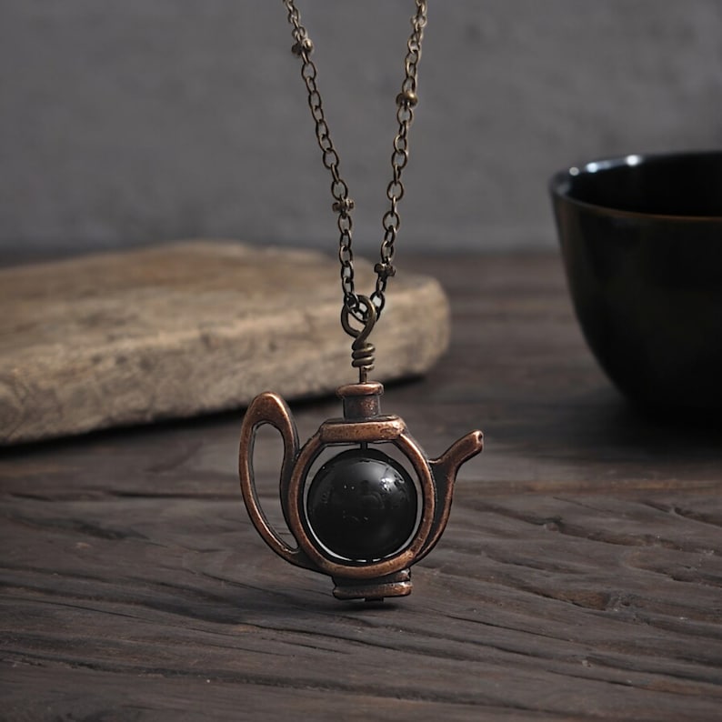 May include: A copper-toned necklace with a teapot pendant. The teapot is open and has a black gemstone inside. The necklace is on a wooden surface.