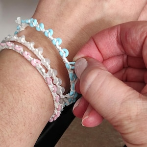 May include: Three layered bracelets with white string and small blue and pink beads. The top bracelet is blue, the middle bracelet is white, and the bottom bracelet is pink.
