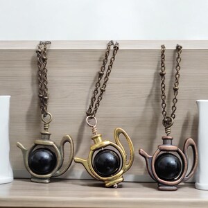 Pode incluir: Três colares dourados com um pingente de chaleira. Cada chaleira tem uma bola preta dentro que gira. As chaleiras estão em diferentes acabamentos: latão antigo, ouro e cobre.