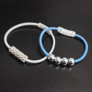 May include: Two hair ties. One is white with a cluster of small silver beads. The other is light blue with three silver beads. Both have a white knotted section.