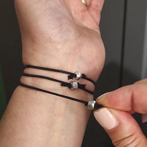 May include: Black cord bracelets with silver beads. The bracelets are wrapped around a wrist. The beads have a textured surface. The bracelets are adjustable.