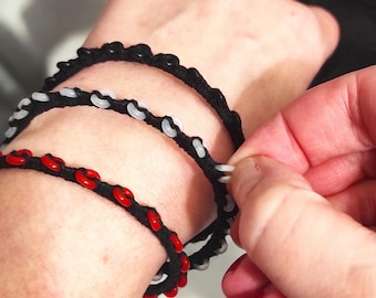 May include: Three black rope bracelets with colorful accents are displayed on a wrist. One bracelet features white beads, another has red beads, and the third has black beads. The bracelets are handmade and have a unique, textured design.