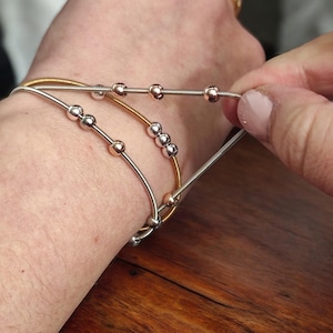 May include: Three silver and gold bracelets with small beads. The bracelets are stacked on a person's wrist.