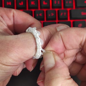 May include: A white beaded ring is being adjusted by a person's fingers. The ring is made of small, round, white beads strung together with white thread. The background shows a red-lit keyboard.