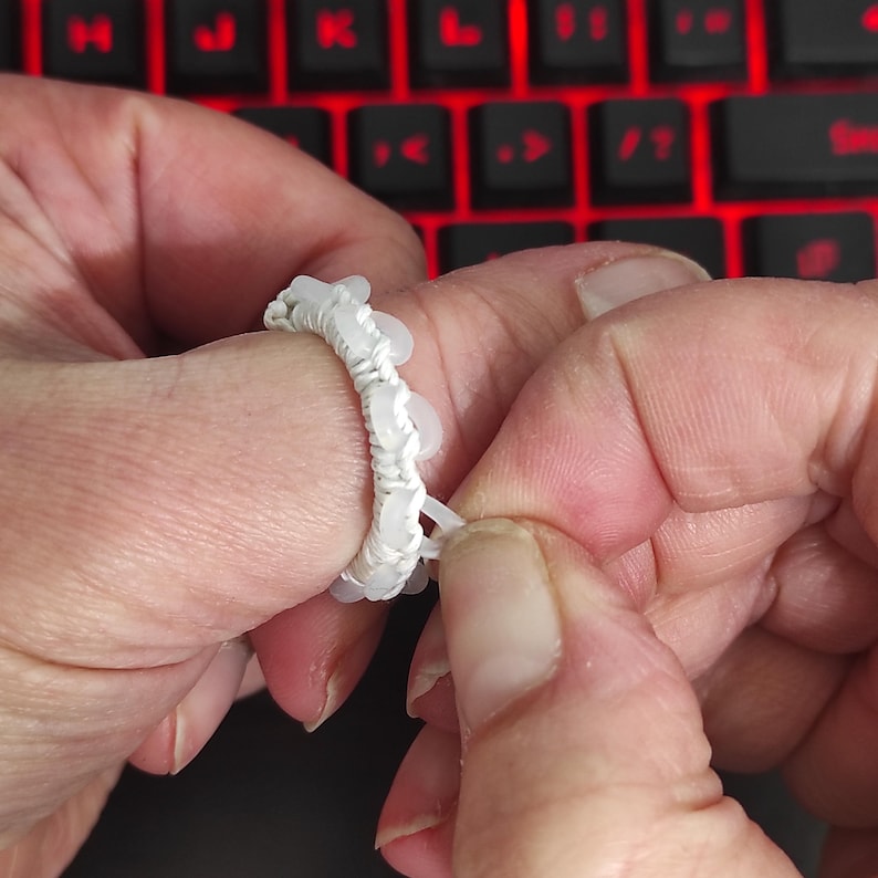 May include: A white beaded ring is being adjusted by a person's fingers. The ring is made of small, round, white beads strung together with white thread. The background shows a red-lit keyboard.