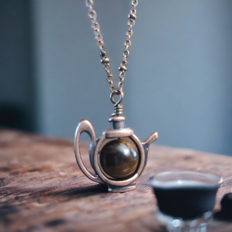 May include: A gold-toned necklace with a teapot pendant featuring a round, brown tiger's eye stone.