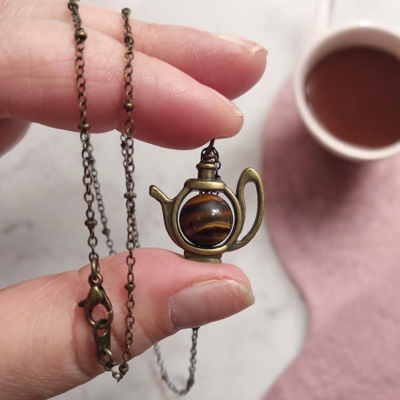 May include: A gold-toned necklace with a teapot pendant featuring a tiger's eye stone. The teapot is detailed with a spout, handle, and a round body.