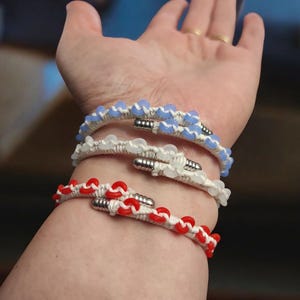 May include: Three handmade bracelets in blue, white, and red. Each bracelet features a woven cord design with decorative beads and a silver-colored clasp. The bracelets are worn on a wrist.