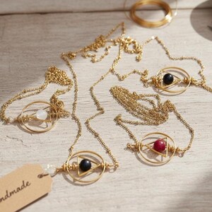 May include: Gold-colored necklaces with geometric pendants. Each necklace features a circular frame with a triangle and a gemstone in the center. Gemstones include white, black, and red. The necklaces are displayed on a light-colored wooden surface.