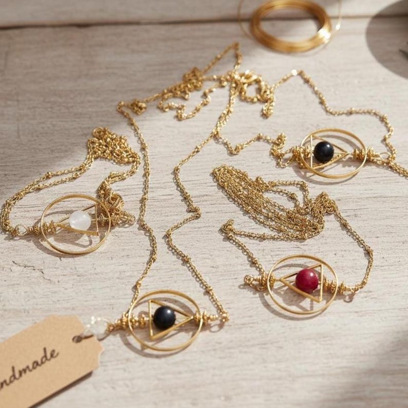 May include: Gold-colored necklaces with geometric pendants. Each necklace features a circular frame with a triangle and a gemstone in the center. Gemstones include white, black, and red. The necklaces are displayed on a light-colored wooden surface.