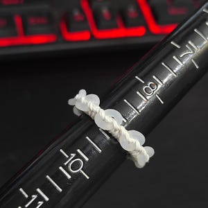 May include: A handmade ring featuring a white cord adorned with small, translucent, circular beads. The ring is displayed on a black ring sizer, with measurements in inches and a red-lit keyboard in the background.