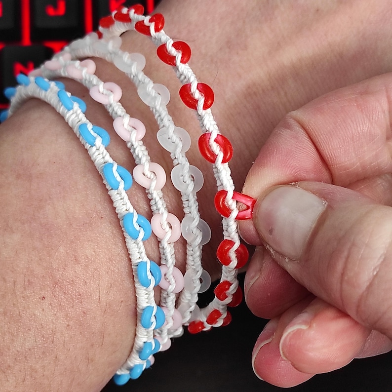 May include: Close-up of several handmade bracelets. Each bracelet is made of white cord and adorned with colorful heart-shaped beads in red, pink, blue, and white. The bracelets are being held by a hand.