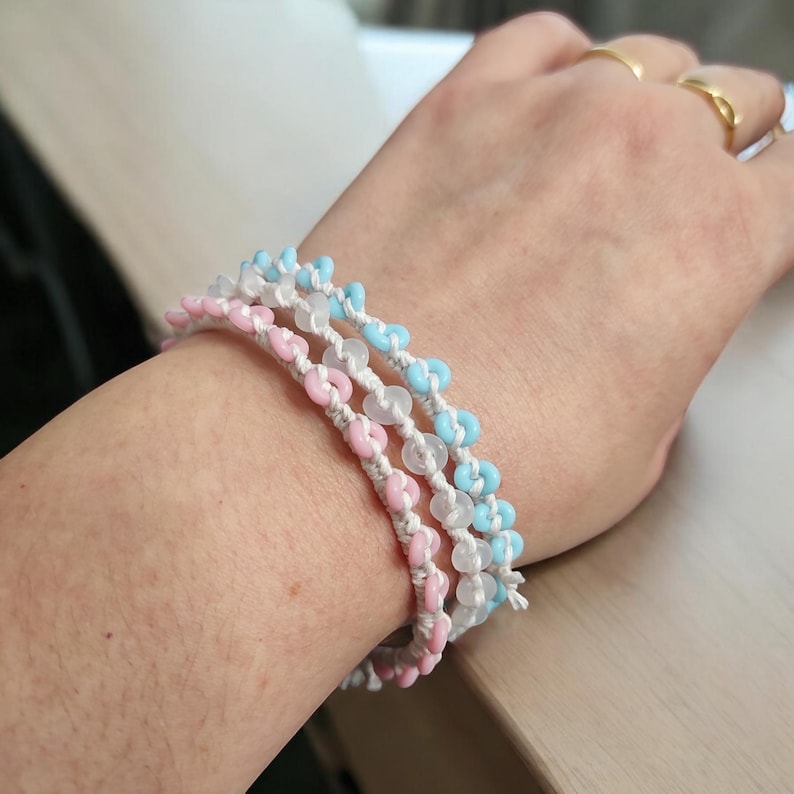 May include: Three handmade bracelets with pink, blue, and clear beads strung on white cord. The bracelets are stacked on top of each other. The beads are small and round, and the cord is tightly woven.