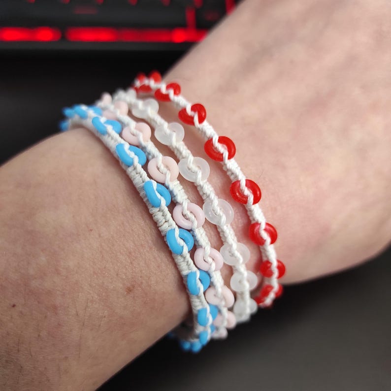 May include: Four handmade bracelets with white cord and colorful beads. The bracelets feature blue, pink, white, and red beads. The beads are strung on a white cord, creating a layered effect. The bracelets are worn on a wrist.