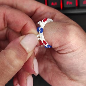 May include: A ring constructed from white cord knotted in a macrame style, featuring small, smooth, circular beads in alternating red and blue colours integrated into the band. The ring is flexible and designed to be worn on a finger.