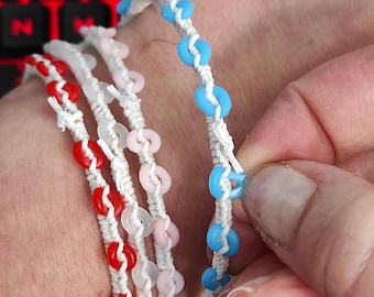 May include: Close-up of several handmade bracelets. Each bracelet features colorful, circular beads strung on white cord. The beads are red, pink, and light blue. The bracelets are displayed on a wrist.
