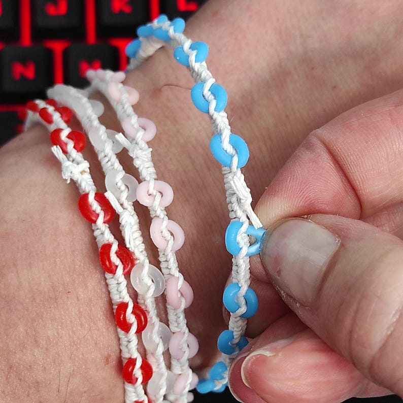May include: Close-up of several handmade bracelets. Each bracelet features colorful, circular beads strung on white cord. The beads are red, pink, and light blue. The bracelets are displayed on a wrist.