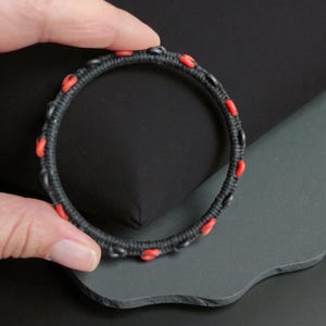 May include: A black bracelet with red accents. The bracelet is made of a woven material and features small, evenly spaced red beads. The design is simple and elegant, suitable for everyday wear or special occasions.